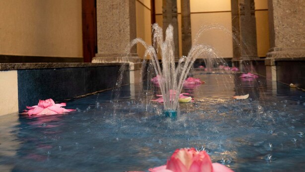 Una fuente con chorros de agua en una piscina, rodeada de columnas de piedra, en un edificio con techos altos y una puerta al fondo.