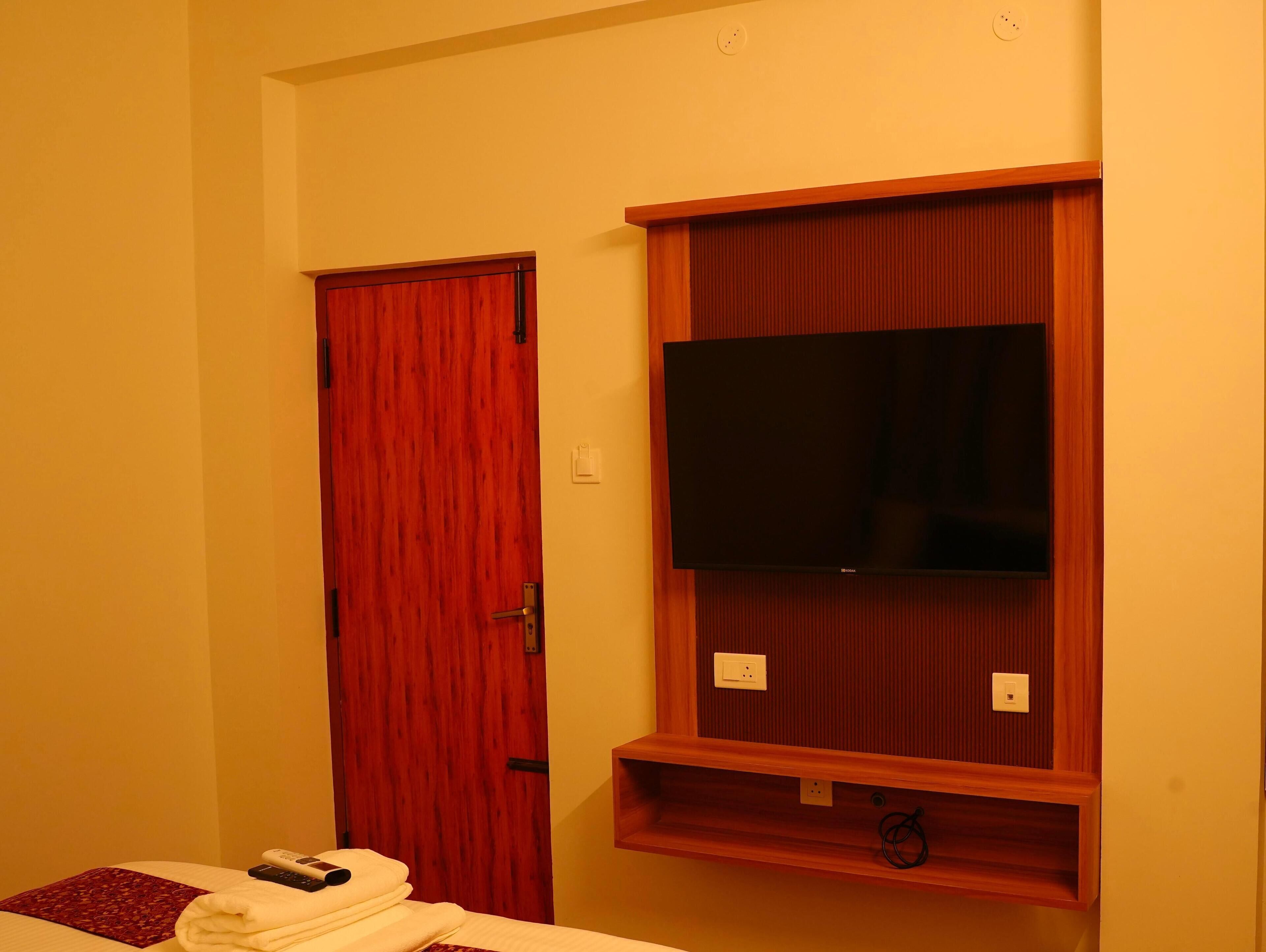 Una habitación de hotel con una puerta de madera, un televisor de pantalla plana montado en un panel de madera, y una cama con un control remoto y toallas.