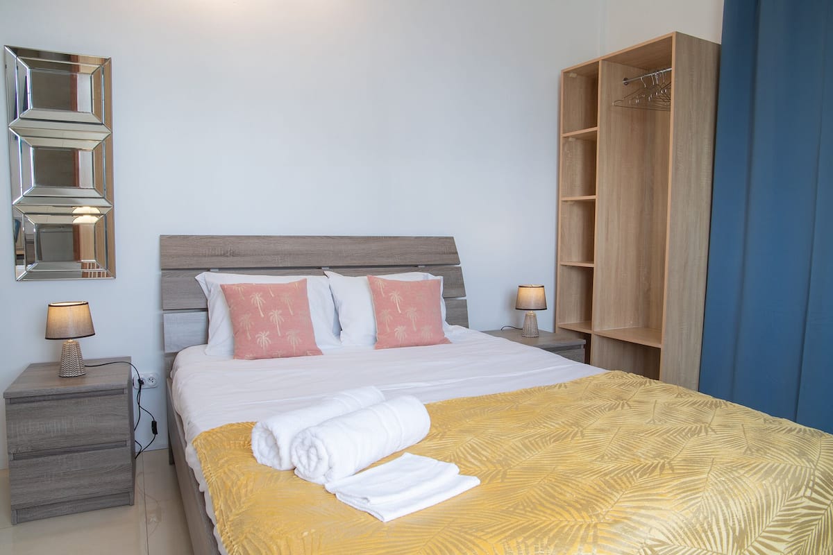 Una cama bien tendida con una colcha amarilla estampada, dos almohadas con diseño de palmera, un cabecero de madera, lámparas de noche, un espejo y un armario de madera.