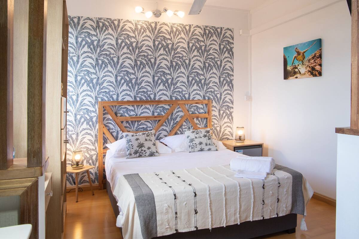 Un dormitorio con una cama, cabecera de madera, papel tapiz estampado, mesita de noche, lámpara y un cuadro en la pared.
