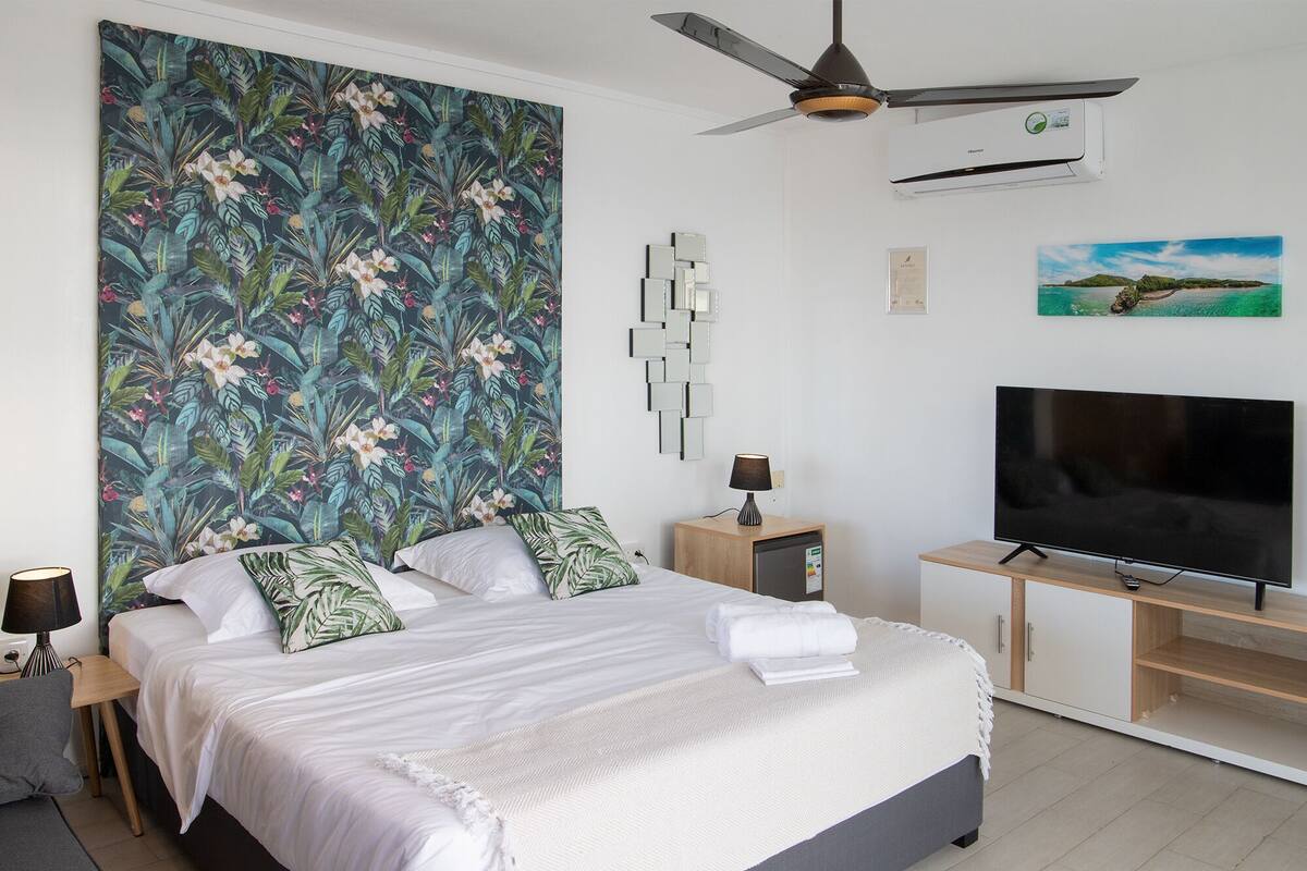 Una habitación de hotel moderna con una cama, un televisor de pantalla plana, un ventilador de techo y un papel tapiz con motivos tropicales.