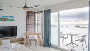 A hotel room with a bed, a desk with a TV, a chair, a balcony with a table and chairs, and a view of the sea. - Casa Maria (Pointe aux Sables)