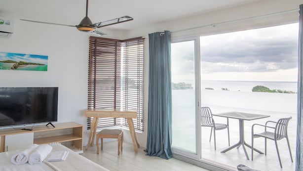 Een hotelkamer met een bed, een bureau met een televisie, een stoel, een balkon met een tafel en stoelen, en uitzicht op zee.