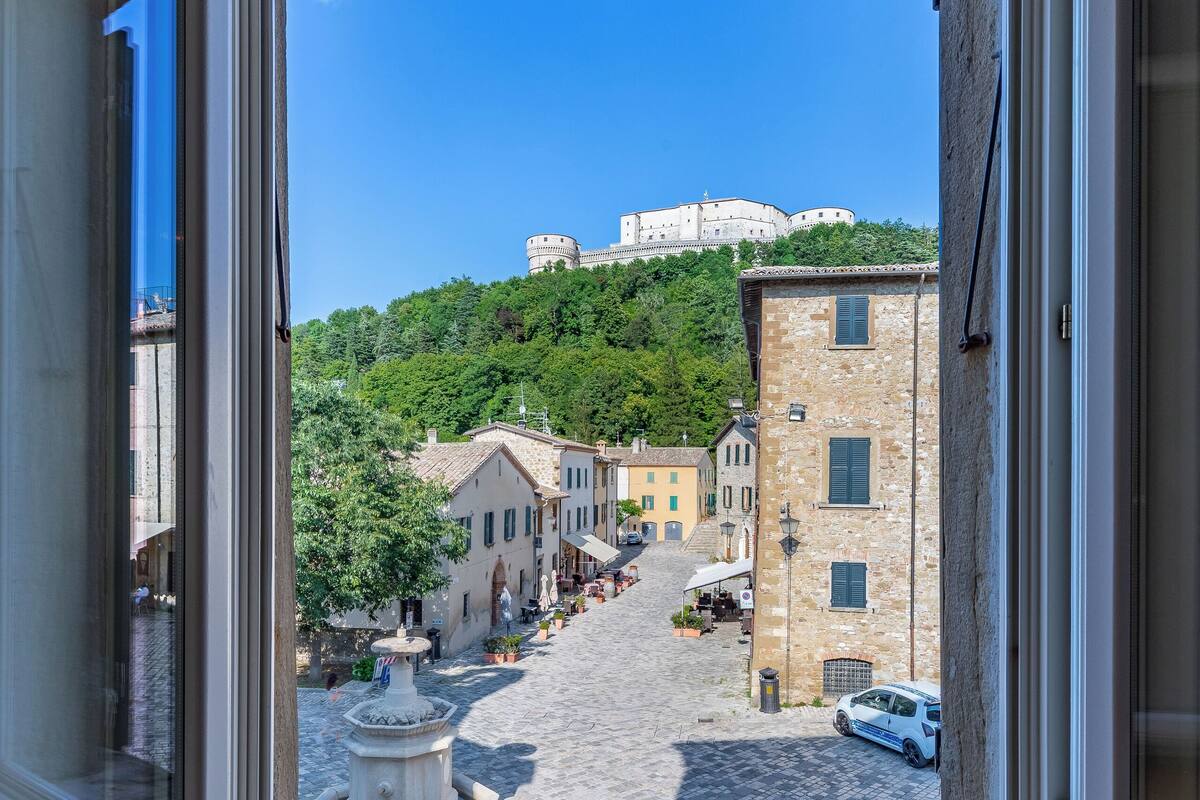 A view from a window overlooking a cobblestone street with buildings, a fountain, and a hill with a castle-like structure in the distance.
