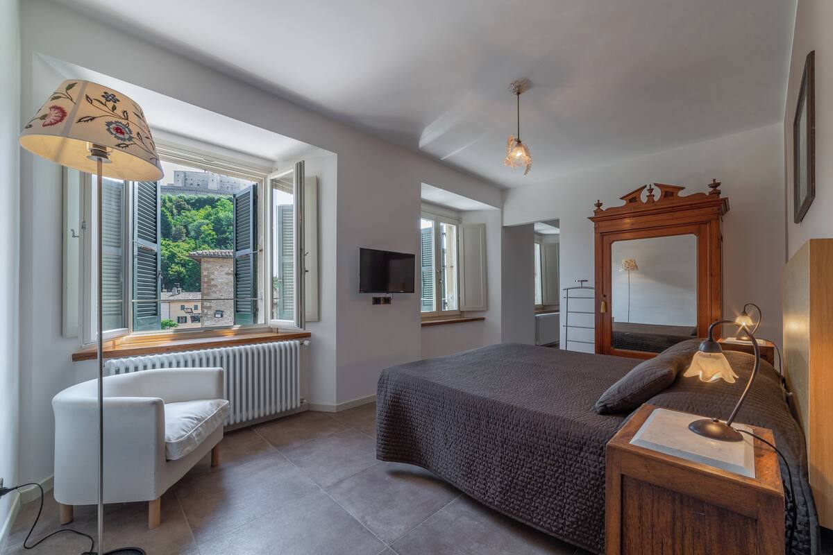 A bedroom with a large bed, a white chair, a floor lamp, a wooden nightstand, and a TV.