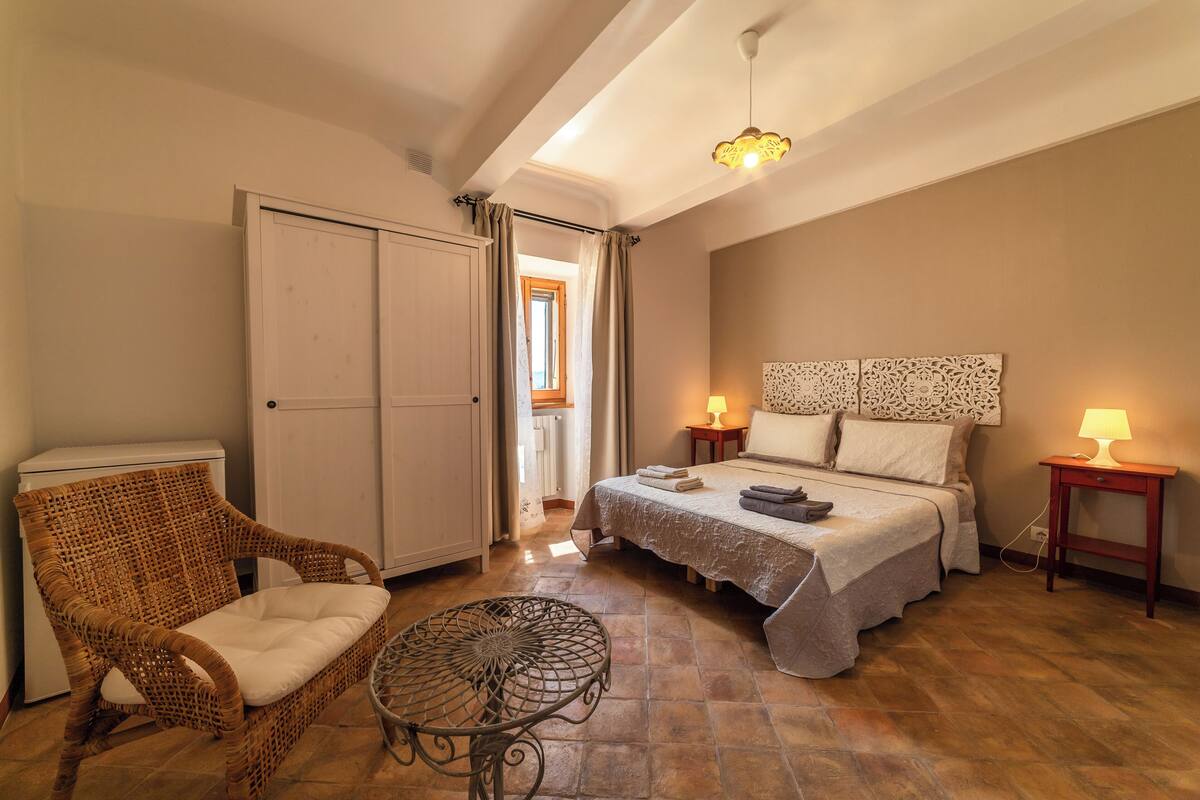 A bedroom with a bed, a wicker chair, a small table, a wardrobe, and a nightstand with a lamp.