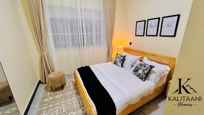 Une chambre à coucher bien rangée, avec un lit, une lampe de chevet et un petit tabouret. Trois tableaux encadrés sont accrochés au mur.