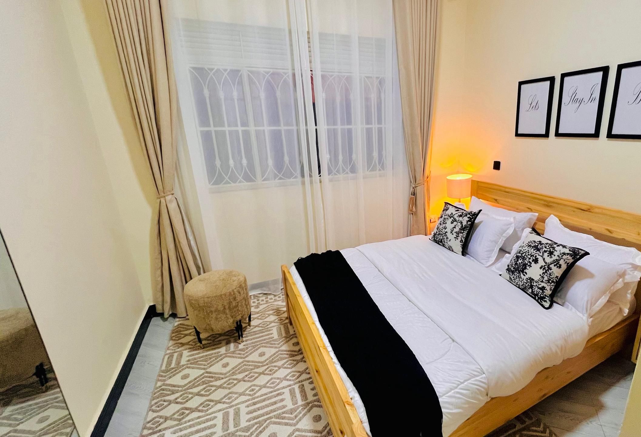 A neatly arranged bedroom with a bed, a bedside lamp, and a small stool. There are three framed pictures on the wall.