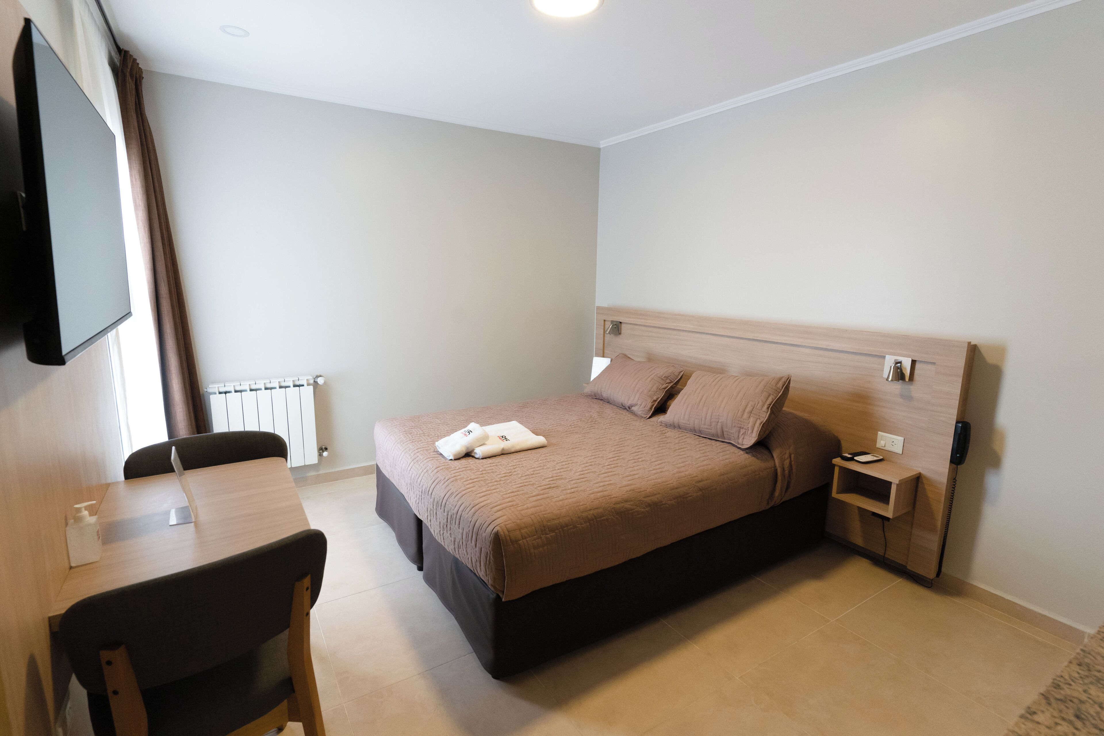 A hotel room with a bed, bedside table, lamp, and a desk with a chair.