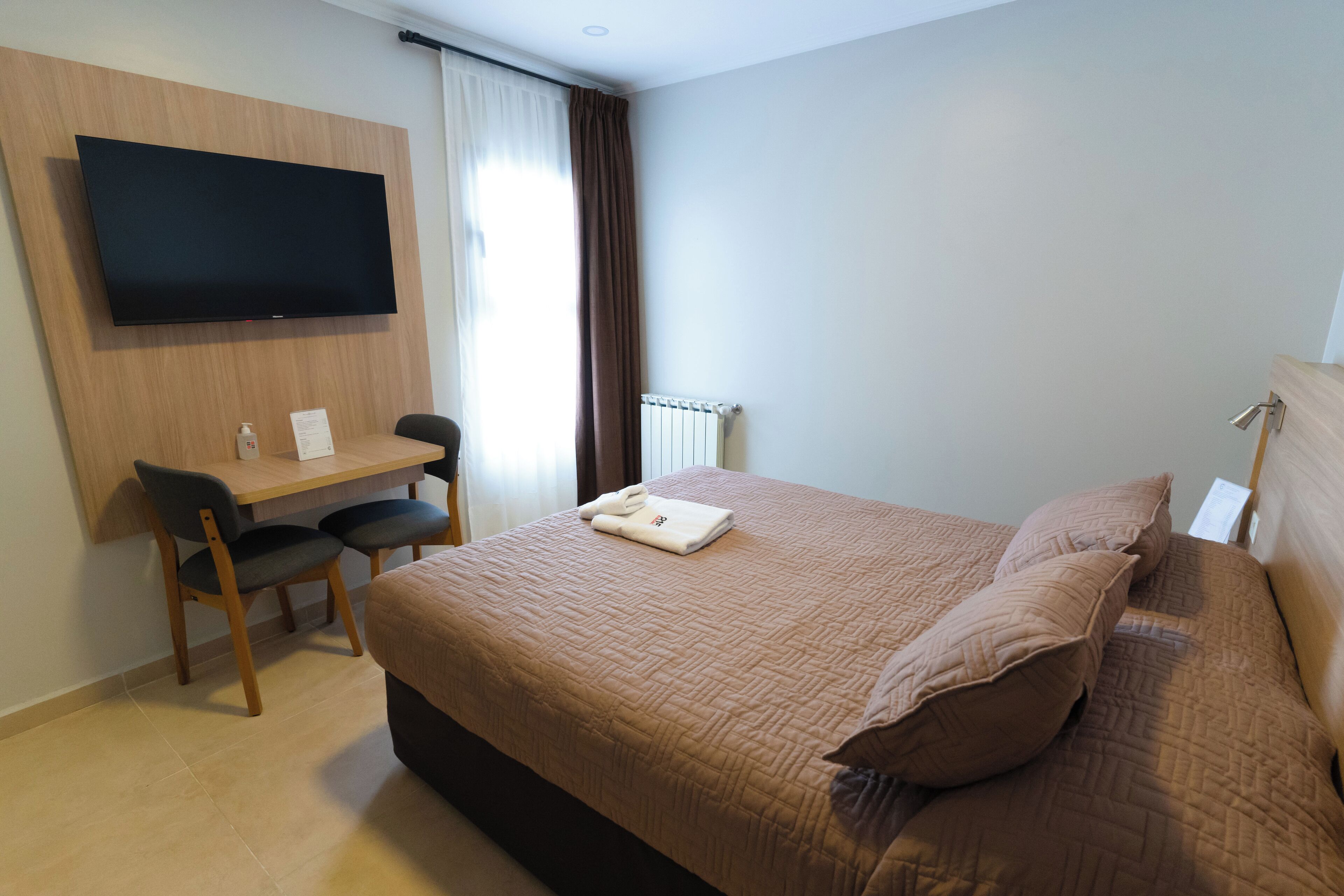 A hotel room with a bed, a desk, a chair, a flat-screen TV mounted on the wall, and a window with curtains.