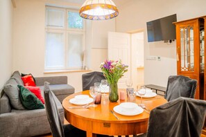 A dining area with a round wooden table set for four, surrounded by chairs. A vase with flowers is on the table, and there is a television mounted on the wall.