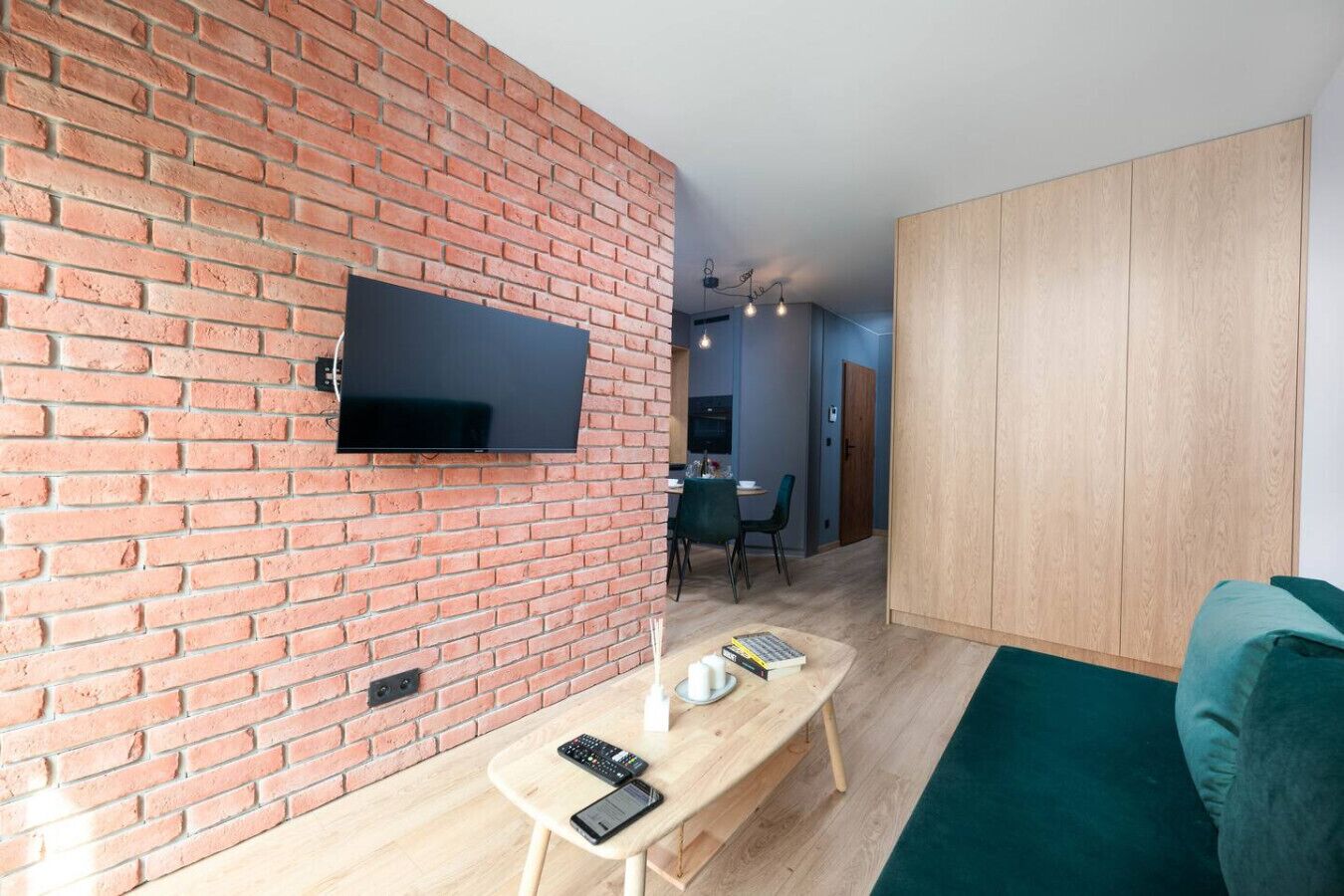 A modern living space with a brick wall, a flat-screen TV mounted above a wooden table, a green sofa, and a wooden wardrobe.