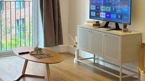 Ein modernes Wohnzimmer mit einem Flachbildfernseher, einem Holztisch, einer Essecke mit Schüssel und Besteck sowie Blick auf ein Backsteingebäude durch ein großes Fenster.