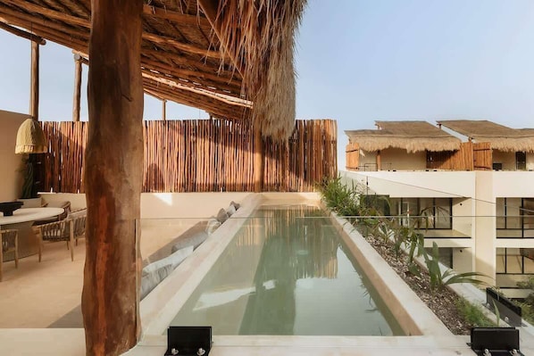 A pool area with a wooden deck, a thatched roof structure, and a modern building with large windows.