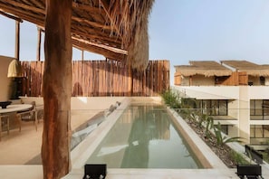 A pool area with a wooden deck, a thatched roof structure, and a modern building with large windows.