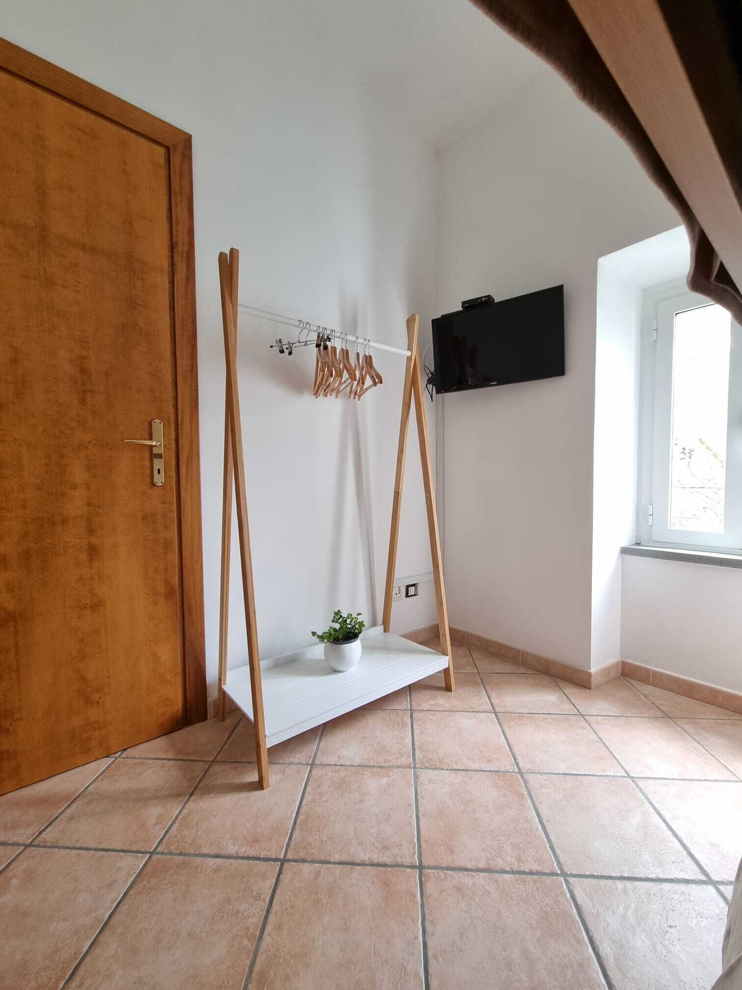 A room with a wooden door, a white shelf with a plant, a clothes rack, and a TV mounted on the wall.