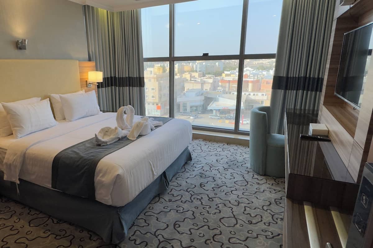 A hotel room with a large bed, a city view through the window, a flat-screen TV, and a desk with a telephone.