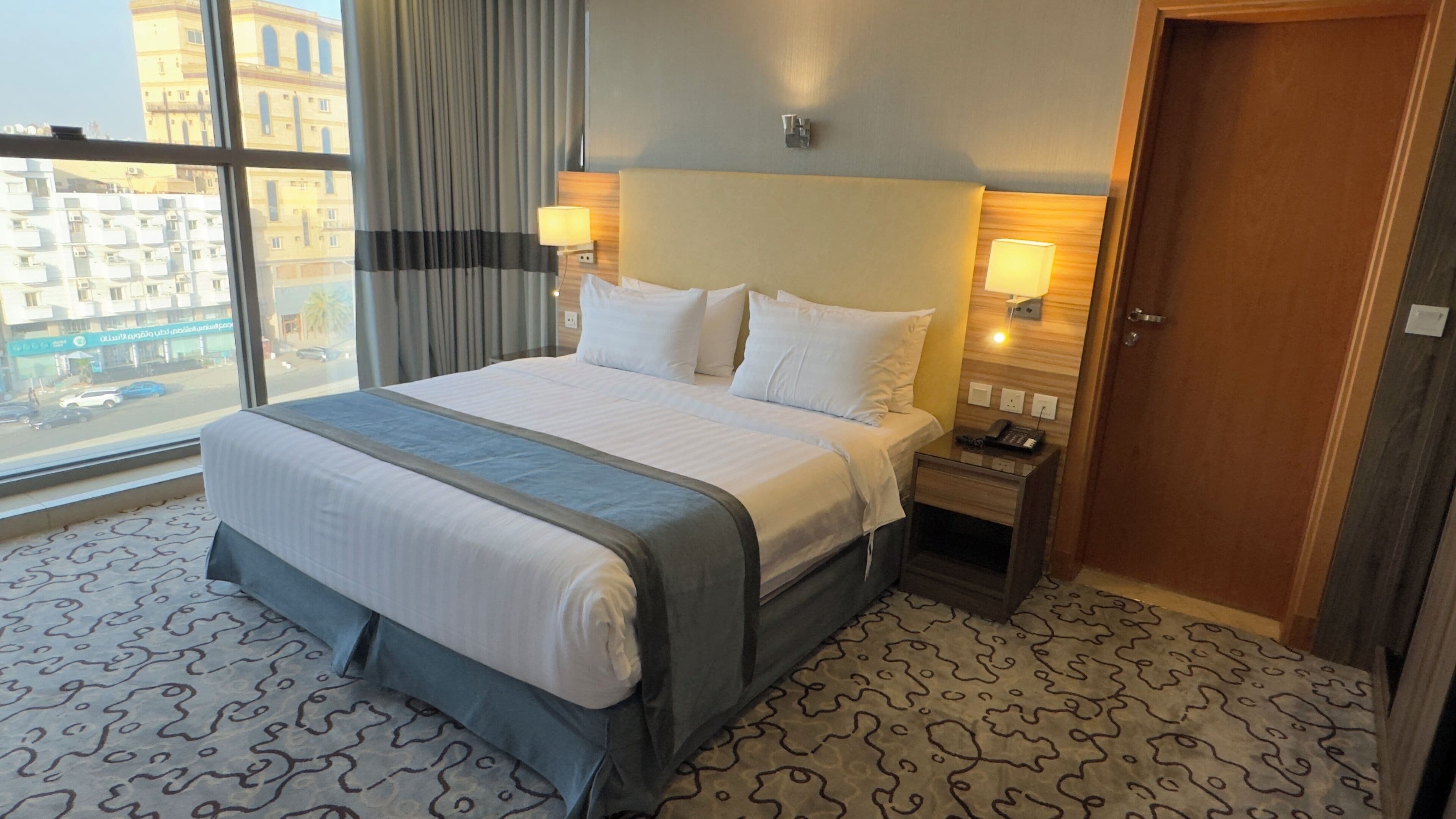A hotel room with a large bed, bedside lamps, a nightstand, and a view of the city through large windows.