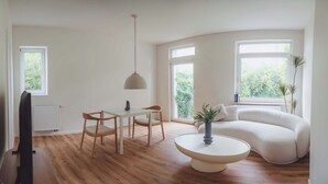A modern living room with a wooden floor, a white sofa, a dining table with chairs, a round coffee table, and large windows offering a view of greenery. - Boutique Suite by the Park w Parking (Celle)