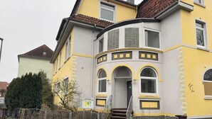 A two-story building with a curved bay window, a staircase leading to the entrance, and a metal fence in front. - Boutique Suite by the Park w Parking (Celle)