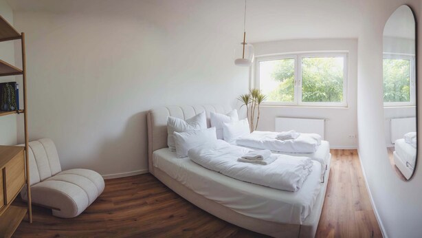 A modern bedroom with a curved bed, a wooden sideboard, a large window with a view of greenery, and a mirror on the wall. - Boutique Suite by the Park w Parking (Celle)