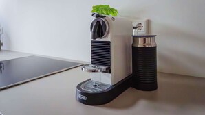 Eine Kaffeemaschine mit schwarzem Fuß und weißer Oberseite, auf einer weißen Arbeitsplatte neben einem schwarzen Gegenstand.