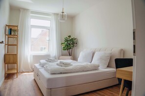 A modern bedroom with a large bed, a wooden shelf, a potted plant, and a window with curtains.
