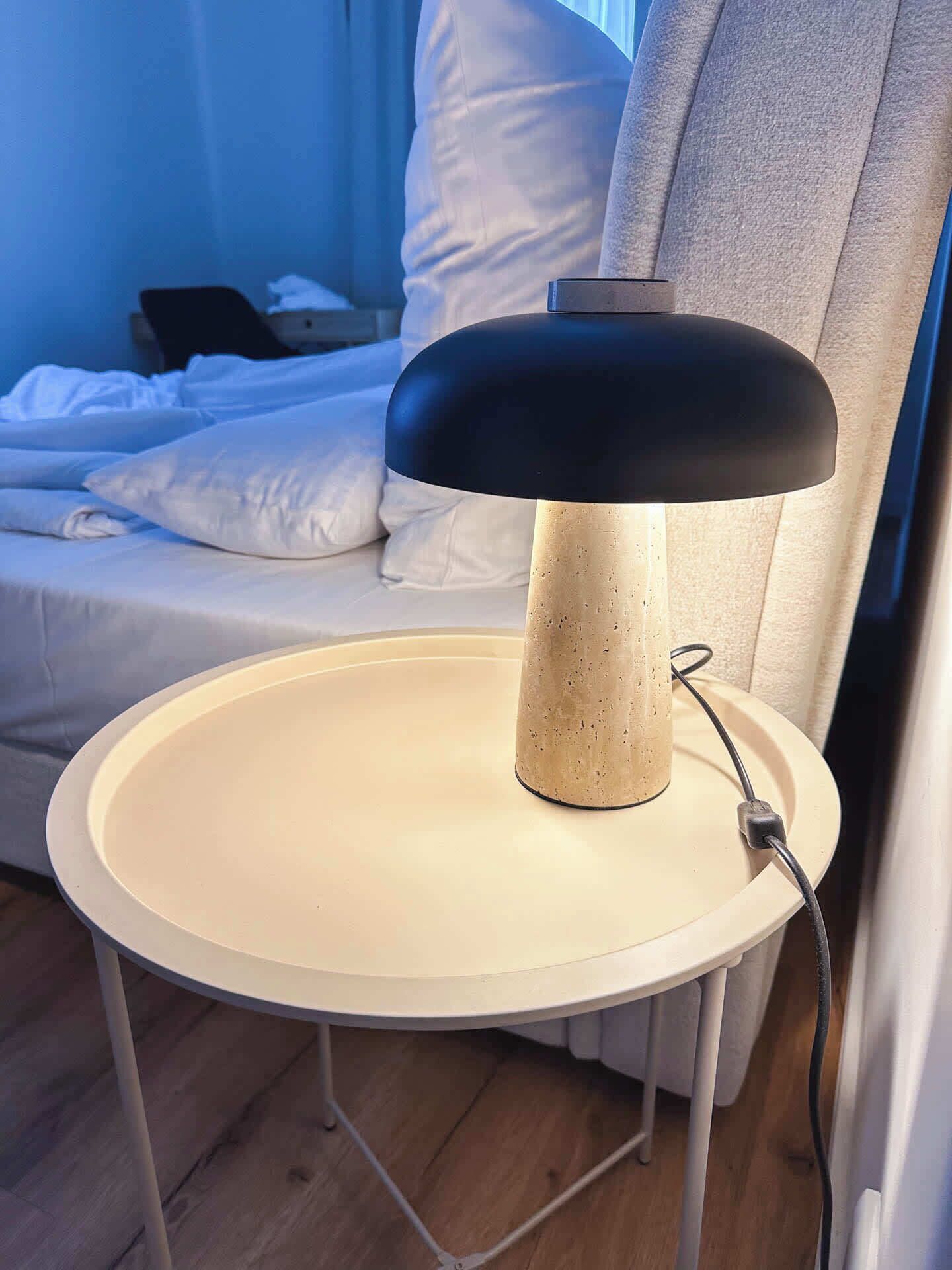 A bedside table with a lamp, a bed, and a window with curtains.