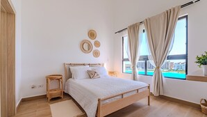 Une chambre avec un lit en bois, une petite table de chevet, une grande fenêtre avec des rideaux et une vue sur des immeubles et une piscine.