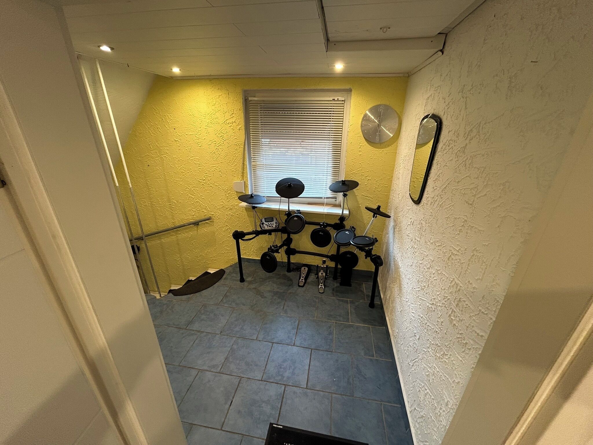 A room with a drum set, a window with blinds, and a mirror on the wall.