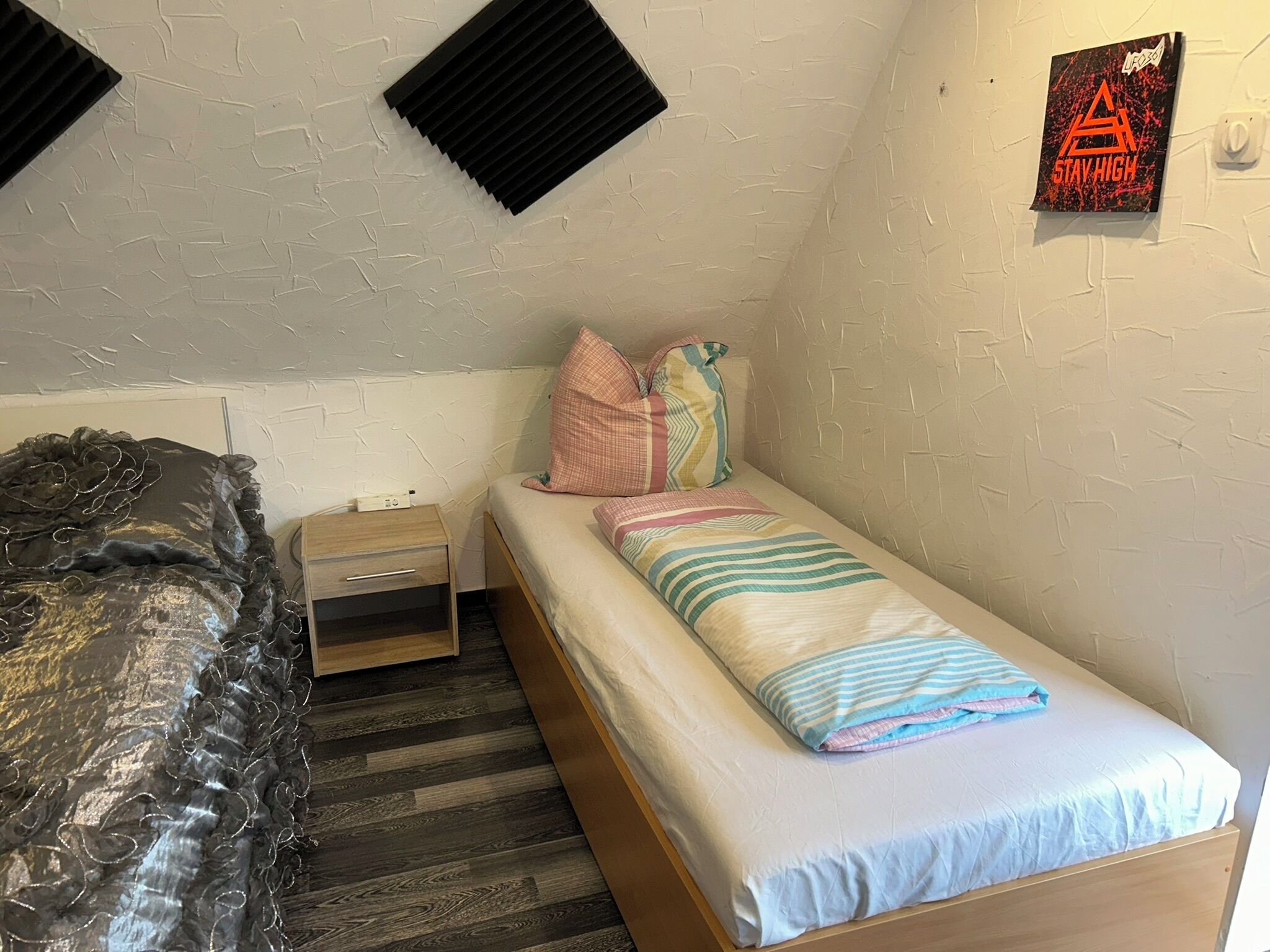 A small, single-bed room with a wooden bed, a nightstand, and a wall-mounted shelf.