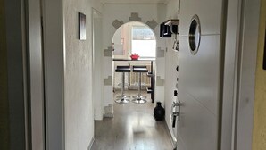 Un couloir mène à une cuisine où l’on aperçoit une casserole rouge sur le plan de travail, une fenêtre et une porte sur le côté gauche.