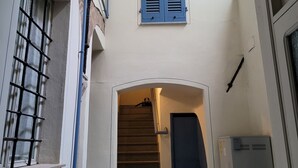 Eine schmale Treppe führt zu einer dunklen Tür, begleitet von weißen Wänden und einem Fenster mit blauen Rollläden.