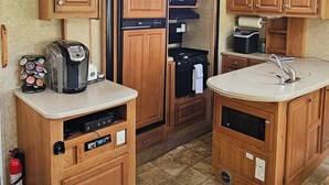 A compact RV kitchen with wooden cabinets, a built-in stove, and a small countertop with a coffee maker. - A&J Country RV Getaway (Gilman)
