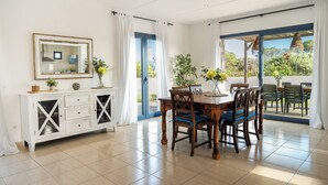 A dining area with a wooden table and chairs, a white cabinet with drawers, and a large window with curtains. - Elegant and modern detached villa with swimming pool (Santanyí)