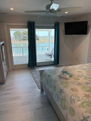 A bedroom with a bed, a TV, a ceiling fan, a view of a lake, and a balcony with a chair.