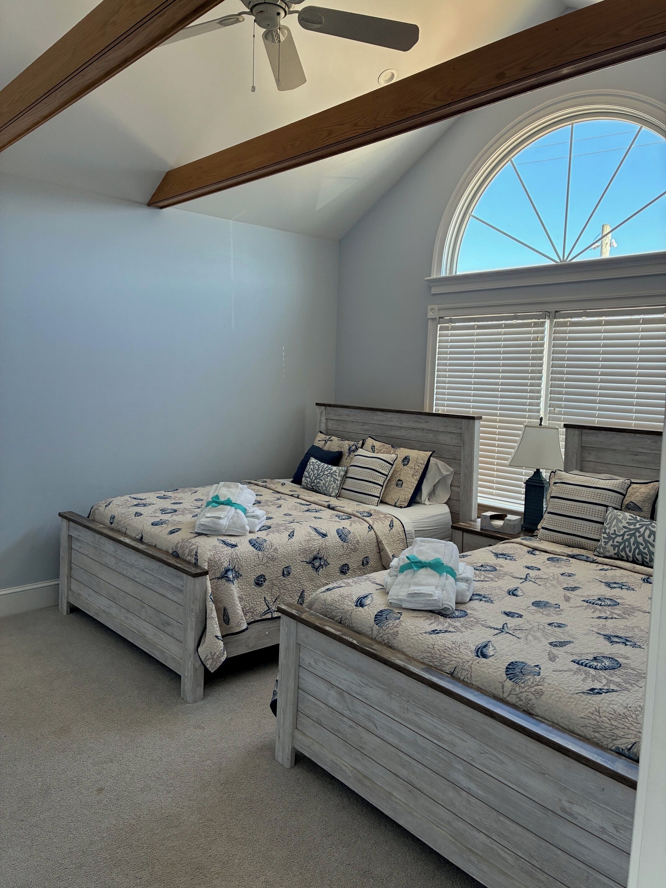 A bedroom with two wooden beds, a ceiling fan, a large arched window, and a lamp on a bedside table.
