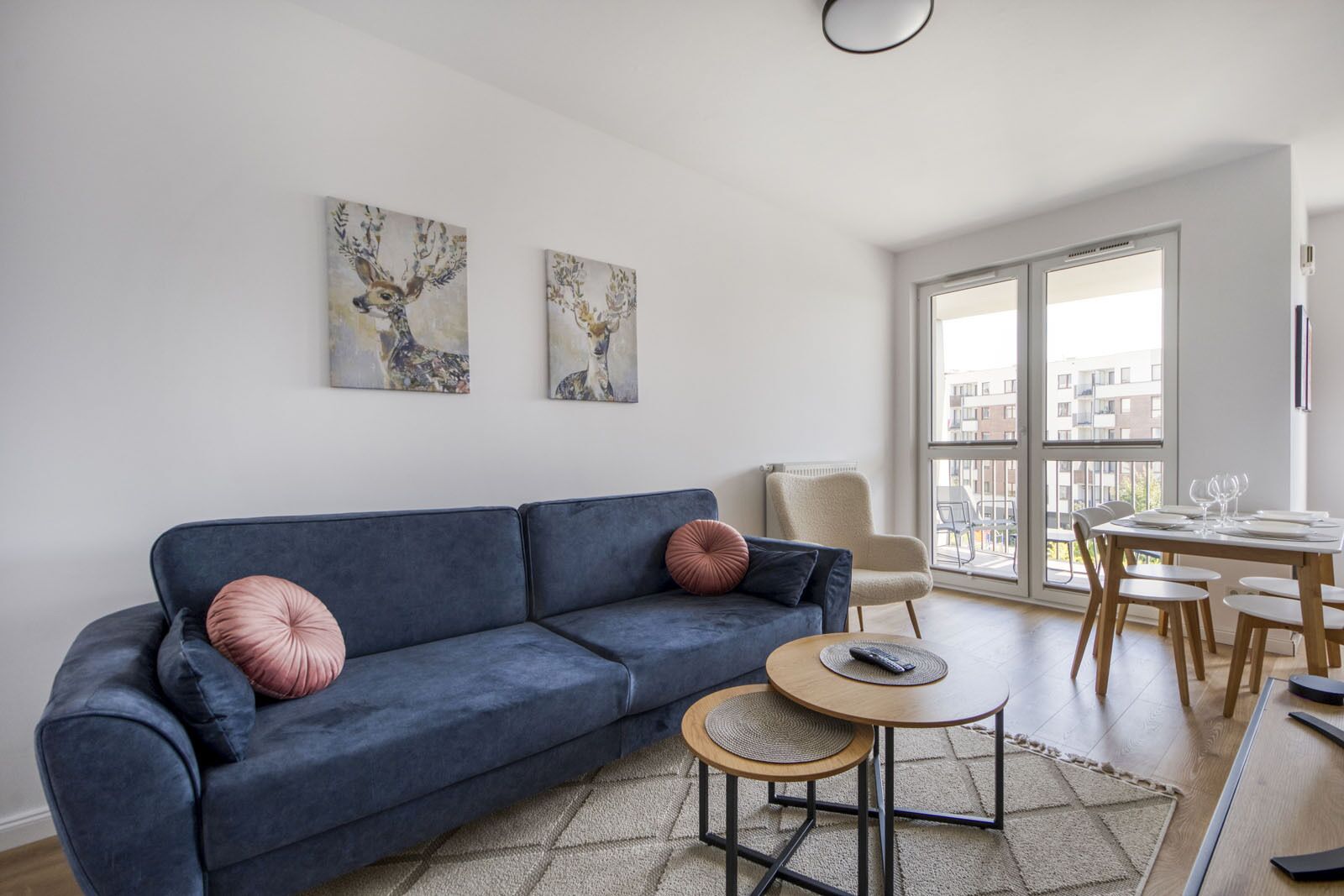 A modern living room with a dark blue sofa, two decorative wall art pieces, a small round table with a remote control, and a dining area with a wooden table and chairs.