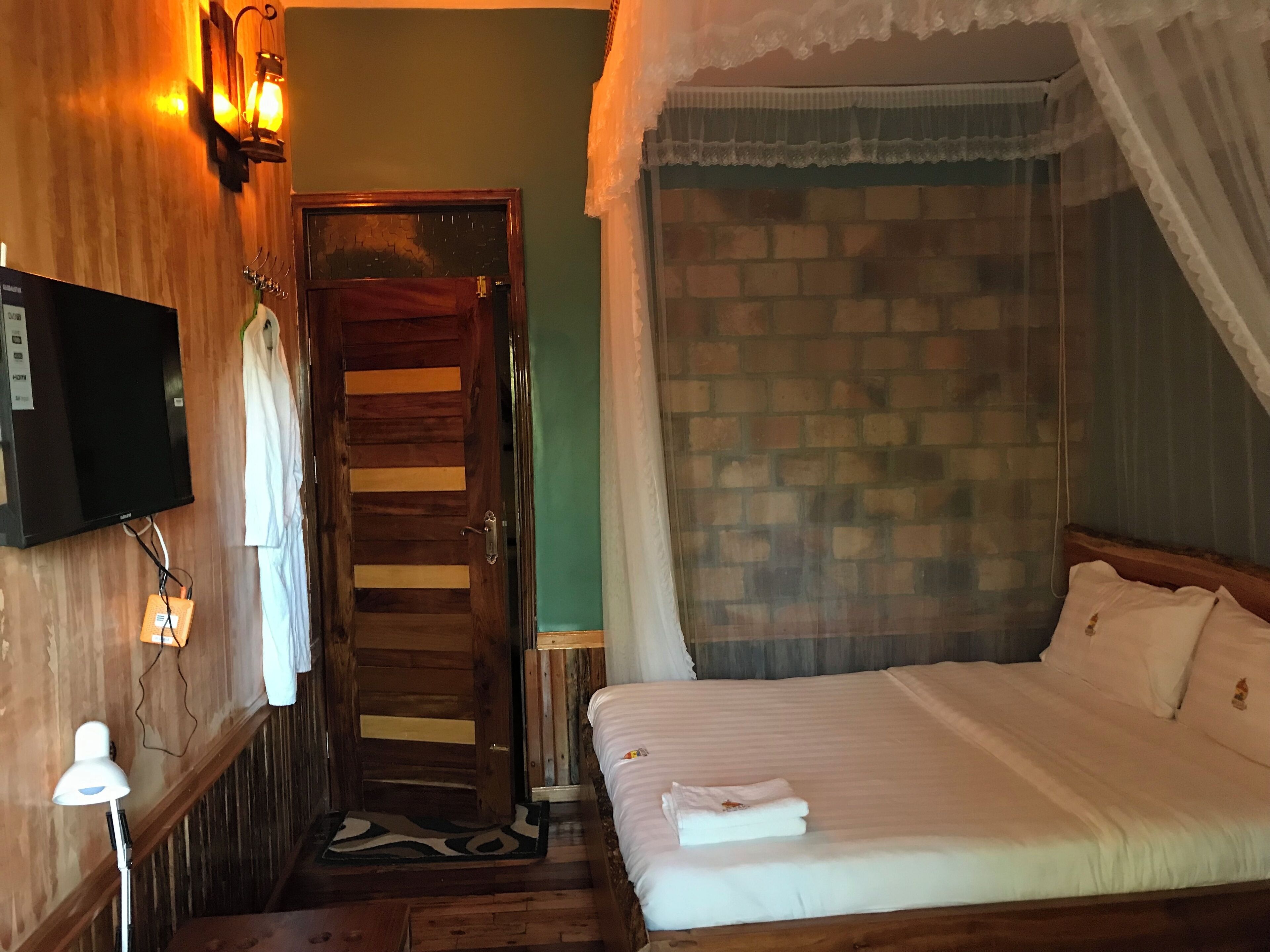 A room with a wooden door, a bed with white bedding, a TV mounted on the wall, and a brick wall.
