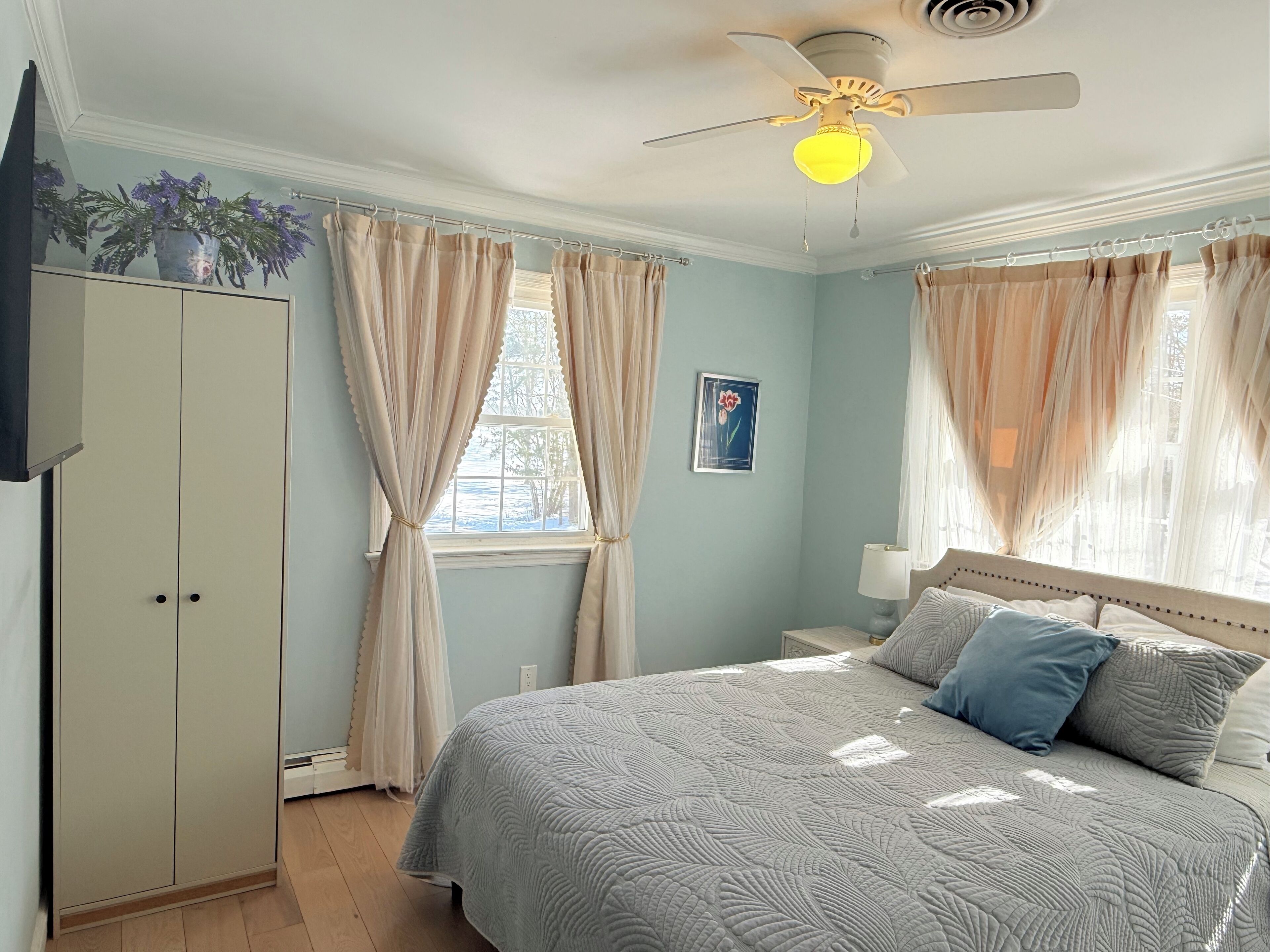 Ein Schlafzimmer mit Bett, Deckenventilator, Fenster mit Vorhängen, Nachttisch und einem an der Wand montierten Fernseher.