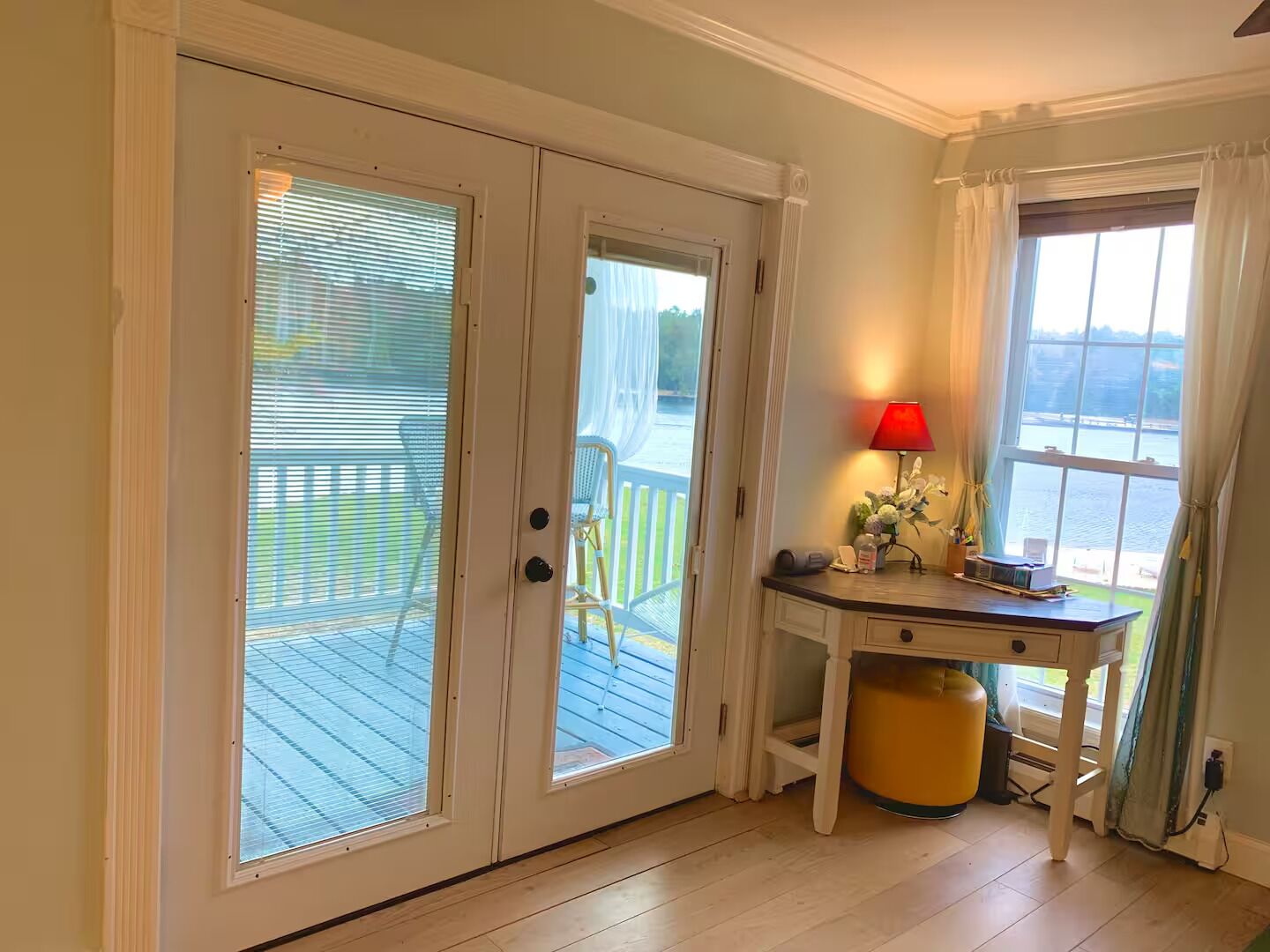 A room with a wooden floor, a desk with a lamp, a trash can, and a view of a deck and water outside.