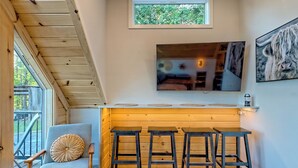 A cozy cabin interior with a wooden ceiling, a dining area with four stools, a large TV mounted above a wooden counter, and a window with a view of trees.