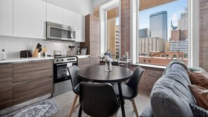 Une cuisine moderne avec des armoires blanches, un four à micro-ondes en acier inoxydable et une cuisinière. On y trouve un coin repas avec une table ronde et des chaises, ainsi qu’une grande fenêtre offrant une vue sur la ville.