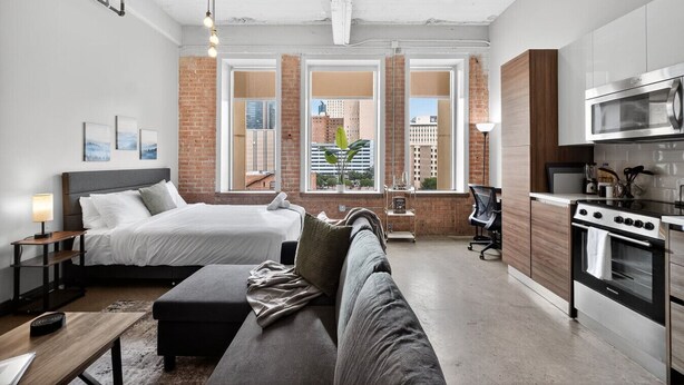 A modern hotel room with a large bed, a sofa, a kitchen area with a stove and microwave, and large windows with a city view. - Prime Downtown Dallas | Pool + Theater + Game Room (Dallas)