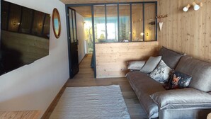 Ein gemütliches Wohnzimmer mit einem großen Flachbildfernseher, einer Holzbalkendecke, einer Couch mit gemusterten Kissen und Blick durch eine Glastür auf einen Außenbereich.