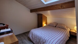Een slaapkamer met dakraam, een bed met gestreept beddengoed, een houten nachtkastje met een lamp en een klein tafeltje met boeken.