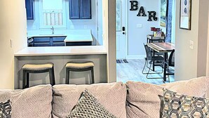 A modern kitchen with dark cabinets and a white countertop, a dining area with a bar sign, and a cozy seating area with patterned cushions. - Charming Coastal Cottage Near Beach & Downtown (Wilmington)