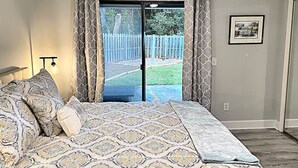 A bedroom with a bed, patterned bedding, a nightstand, a lamp, and a view of a backyard through a sliding glass door. - Charming Coastal Cottage Near Beach & Downtown (Wilmington)