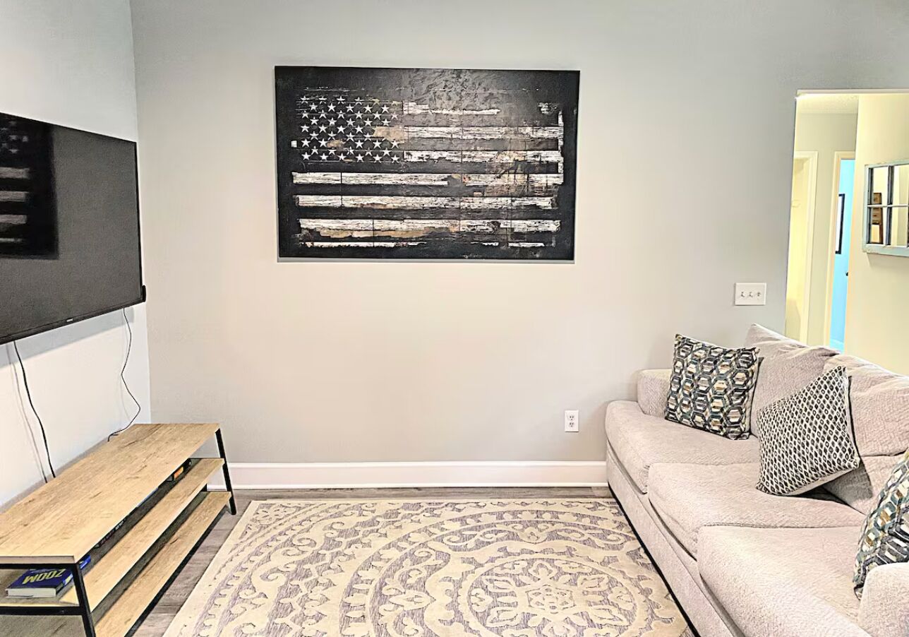 Una sala de estar con una televisión de pantalla plana, una mesa de centro de madera, un sofá beige con cojines estampados y un cuadro grande con una bandera estadounidense.