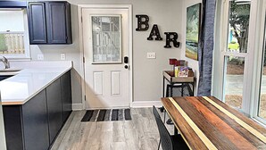 A modern kitchen with dark cabinets, a wooden dining table, and a 'BAR' sign on the wall. - Charming Coastal Cottage Near Beach & Downtown (Wilmington)